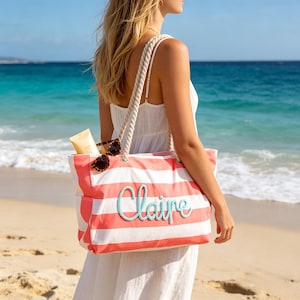 Puede incluir: Un bolso de lona a rayas color coral y blanco con asas de cuerda gruesa y el nombre Claire bordado en letra cursiva azul claro en la parte delantera. El bolso contiene unas gafas de sol de carey y un tubo de protector solar beige.