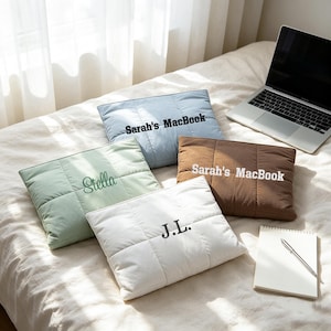 May include: Four rectangular quilted laptop sleeves in light blue, sage green, brown, and white. The blue sleeve reads Sarah's MacBook, the green reads Stella, the brown reads Sarah's MacBook, and the white sleeve features the initials J.L. in black.