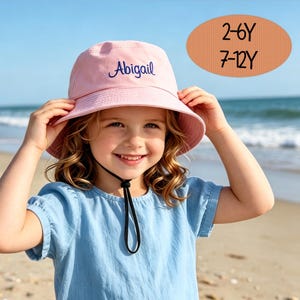 Peut inclure: Un chapeau bob rose clair avec le prénom Abigail brodé en écriture cursive bleu foncé. Le chapeau comprend une mentonnière noire et un graphique ovale affichant le texte 2-6Y et 7-12Y, indiquant les tranches d'âge disponibles pour cet article.