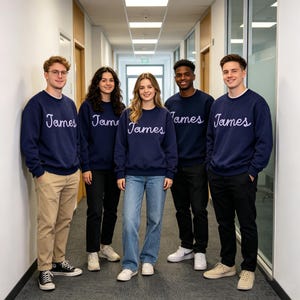 May include: Five individuals standing in a hallway wearing matching navy blue crewneck sweatshirts. Each sweatshirt features the name James embroidered in a light purple cursive script across the chest. The group is dressed in casual pants and sneakers.