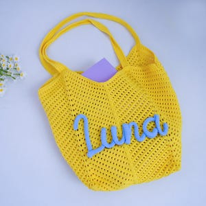 May include: A bright yellow crochet tote bag featuring an open-knit mesh pattern and two shoulder straps. The front of the bag is decorated with the name Luna written in light blue, textured, cursive yarn lettering, with a purple card inside.