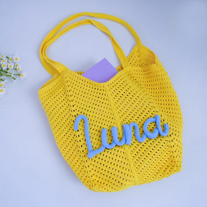 May include: A bright yellow crochet tote bag featuring an open-knit mesh pattern and two shoulder straps. The front of the bag is decorated with the name Luna written in light blue, textured, cursive yarn lettering, with a purple card inside.