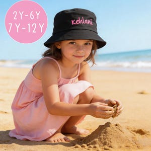 May include: A black bucket hat featuring the name Kehlani embroidered in pink script on the front. A pink circular graphic in the upper left corner displays the text 2Y-6Y and 7Y-12Y, indicating the available age ranges for this headwear item.