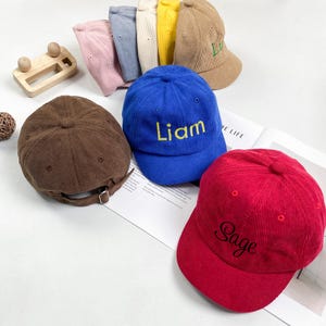 Puede incluir: Una colección de gorras de béisbol de pana en varios colores, incluyendo rosa, azul, amarillo, beige, marrón y rojo. La gorra azul tiene el nombre Liam bordado en amarillo y la roja tiene el nombre Sage bordado en negro.