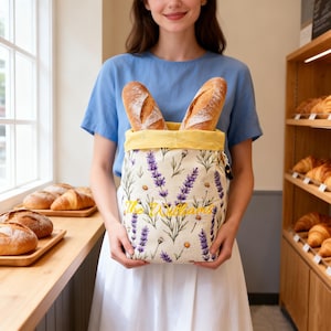 Puede incluir: Una bolsa de tela para pan con fondo crema, estampado floral de lavanda y margaritas, forro interior amarillo y el texto bordado The Williams en amarillo. Dos barras de pan crujientes están colocadas dentro de la parte superior abierta.