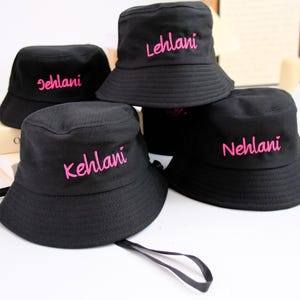 Puede incluir: Cuatro sombreros de pescador negros con texto bordado en hilo rosa brillante. Cada sombrero muestra un nombre único: Lehlani, Kehlani, Nehlani y un nombre parcialmente visible. Los sombreros tienen un ala ancha inclinada y copa plana.