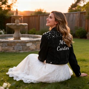 Puede incluir: Una chaqueta vaquera negra con adornos de perlas blancas en los hombros y el texto "Mrs. Carter" en letra cursiva blanca en la espalda, sobre un vestido largo de encaje blanco. La chaqueta tiene cierre de botones y mangas largas.