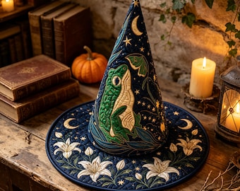 May include: A dark blue quilted witch hat featuring an embroidered green frog, white lily flowers, golden crescent moons, and stars. The conical hat has a wide, flat brim and intricate stitching throughout the fabric, showcasing a celestial design.