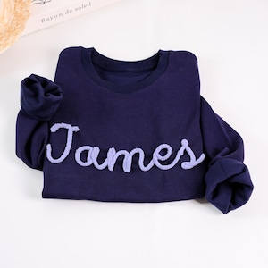 May include: A dark navy blue long-sleeve sweatshirt featuring the name James written in a raised, textured light purple script font across the chest. The garment has a classic crew neckline and ribbed cuffs on the sleeves, providing a simple design.