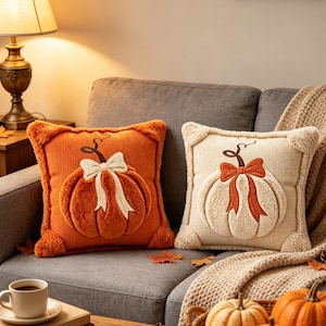 May include: Two square throw pillows featuring a plush, raised pumpkin design with a bow. One pillow is orange with a cream-colored bow, and the other is cream-colored with an orange bow. Both have textured fabric and fuzzy corner accents.