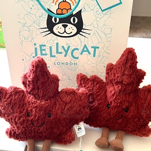 May include: Two plush, burgundy-colored leaf toys with smiling faces and brown legs. The toys are from Jellycat London, as indicated by the tags and shopping bag in the background. The bag features a black cat logo and the brand name.