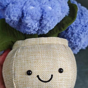 May include: A plush toy featuring a smiling face on a beige fabric body with small brown feet. The toy has a large, fluffy, blue head and green accents. The toy is designed to resemble a plant or flower.