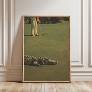 May include: A framed wall art print featuring a golf course scene with a large alligator resting on the green in the foreground, while a golfer in light-coloured trousers stands in the background preparing to strike a golf ball with a club.