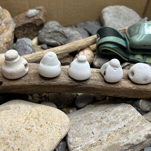 May include: Five small, smooth, white, rounded ceramic objects with holes, arranged in a horizontal row on a piece of weathered driftwood. Each piece has a distinct, slightly irregular shape, resembling worn stones or beads with a matte finish.