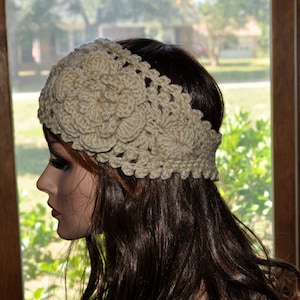 May include: A crocheted headband with a large flower design in a beige color. The headband is worn on a person with long brown hair.
