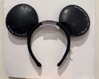 May include: Black headband with two round ears featuring a dark space-themed design with small blue glitter accents. The left ear displays the text A long time ago and the right ear displays the text in a galaxy far, far away in white font.