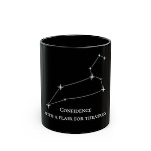May include: Black ceramic mug featuring a white constellation design, with the text "Confidence with a flair for theatrics." The mug is cylindrical and has a glossy finish. The design is a stylized representation of the Leo zodiac sign.