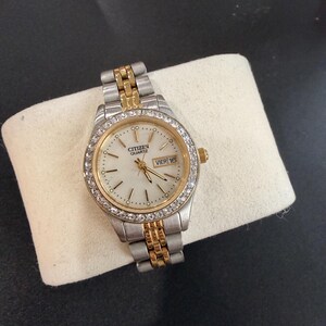 May include: A two-tone Citizen Quartz wristwatch with a silver and gold-colored metal link band. The round watch face features a white dial, gold-tone hands, and a date window displaying VIER 16. The bezel is encrusted with small clear stones.