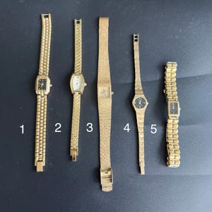 May include: Five gold-tone wristwatches arranged vertically and numbered 1 through 5. Each watch features a metallic link or textured band and a small analogue face with hands. The watches vary in case shape, including rectangular and oval designs.