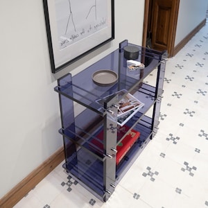 May include: A three-tiered translucent dark blue acrylic shelving unit with silver metal hardware. The top shelf holds a small grey circular tray and a small box. The middle shelf contains magazines, and the bottom shelf holds a red book.