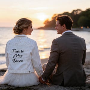 Puede incluir: Una chaqueta vaquera blanca con adornos de perlas en los hombros y el texto negro Future Mrs Breen impreso en la espalda, sobre una falda blanca, junto a una chaqueta de traje gris oscuro, vista desde atrás con un atardecer en la playa de fondo.