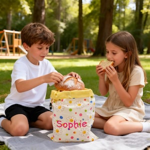 Puede incluir: Una bolsa de tela blanca para pan con un estampado de margaritas amarillas y lunares de colores, con el nombre Sophie impreso en letras rosas en la parte frontal. La bolsa tiene un forro interior amarillo y un cierre de cordón superior.