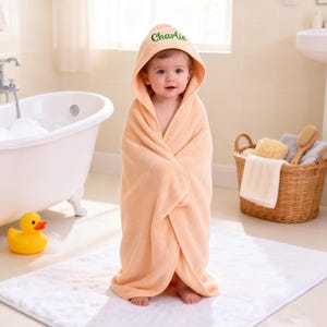 May include: A peach-colored hooded towel featuring the name Charlie embroidered in green script on the hood. The soft, plush fabric wraps around the body, providing full coverage with a pointed hood design that rests comfortably over the head.