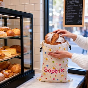 May include: A cream-colored fabric bread bag featuring a repeating pattern of white daisies with yellow centers and colorful polka dots. The name Sophie is printed in pink letters on the front. The bag has a drawstring closure with black toggles.
