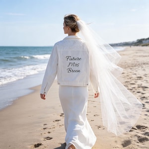 Puede incluir: Una chaqueta vaquera blanca con adornos de perlas en la parte superior de la espalda y el texto negro Future Mrs Breen impreso en el centro. La chaqueta se lleva sobre un vestido largo blanco y un velo de novia de tul blanco transparente.