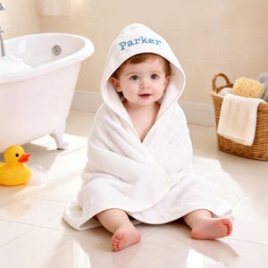 Puede incluir: Una toalla de baño blanca con capucha para bebé que presenta el nombre Parker bordado en hilo azul en la capucha. La toalla está hecha de tela de rizo suave y absorbente, diseñada para envolver al bebé y brindar calidez tras el baño.