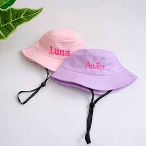 Puede incluir: Dos sombreros de pescador de colores pastel. Uno es rosa claro con el nombre "Luna" bordado en rosa, y el otro es lavanda con "Anika" bordado en rosa. Ambos sombreros tienen correas de barbilla negras.