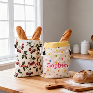 Puede incluir: Dos bolsas de tela para pan con cordones. La bolsa de la izquierda presenta un estampado de cerezas y el texto "Emma's Pantry" en rojo. La bolsa de la derecha tiene un estampado de margaritas blancas y lunares de colores con el nombre "Sophie" bordado en rosa.