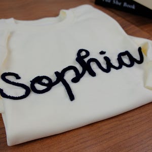 May include: A cream-colored long-sleeve sweatshirt featuring the name Sophia embroidered in a dark navy blue, textured, cursive script across the chest. The garment has a ribbed crew neckline and ribbed cuffs on the sleeves, showcasing a clean design.