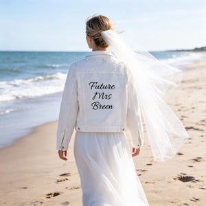 Puede incluir: Una chaqueta vaquera blanca con adornos de perlas en los hombros y la espalda. La parte posterior de la chaqueta muestra el texto negro "Future Mrs Breen" en letra cursiva. Un velo blanco largo y transparente está sujeto al cabello.