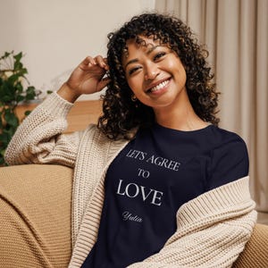 Puede incluir: Camiseta de manga larga azul marino con el texto blanco «LET'S AGREE TO LOVE» y el nombre «Yulia». La camiseta se lleva con una rebeca beige.
