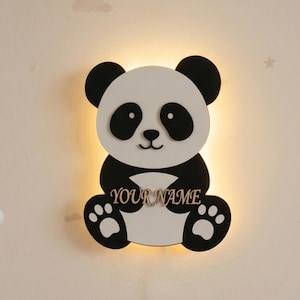 Op de afbeelding: Een panda-vormige wandlamp in zwart en wit. De lamp heeft een uitsparing voor een naam en pootafdrukken op de voeten. De tekst "YOUR NAME" wordt in zilver weergegeven.