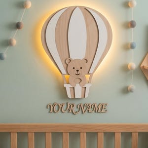 Op de afbeelding: Een houten wandlamp in de vorm van een heteluchtballon met een beer in de mand. De ballon is lichtbruin met witte panelen en de beer is lichtbruin. De tekst "YOUR NAME" staat onder het licht. Het licht staat op een lichtgroene muur.