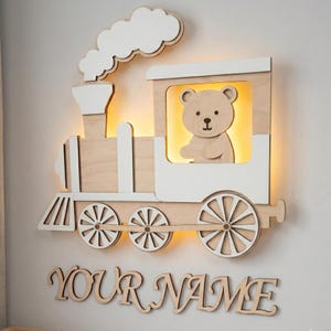 Könnte beinhalten: Eine holzgeschnitzte Wandleuchte in Form einer Lokomotive mit einem Bären als Fahrer. Die Lokomotive ist aus hellem Holz mit weißen Akzenten und einer Rauchwolke gefertigt. Darunter steht "YOUR NAME".