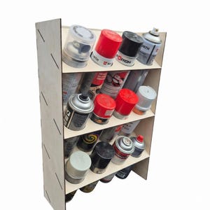 Op de afbeelding: Een lichtgekleurd houten opbergrek met vier schuine planken ontworpen voor spuitbussen. Het rek is gevuld met diverse spuitbussen met verschillende gekleurde doppen, waaronder rood, zwart, wit en zilver, gerangschikt in rijen.