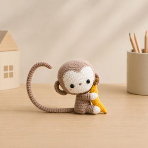 May include: A crocheted monkey doll in shades of brown and white, clutching a yellow banana. The monkey has a long tail and is seated on a wooden surface. The doll is a handmade item.