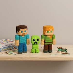 Op de afbeelding: Drie gehaakte Minecraft-karakterfiguren: Steve, een Creeper en Alex. Steve is in blauw, Alex in groen en bruin en de Creeper is groen. Ze worden tentoongesteld op een houten plank met een boek getiteld "BLOCKS & CRAFTS" en knutselspullen.