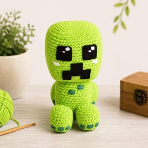 May include: A lime green crocheted plush toy featuring a large square head with black felt eyes and a black mouth. The body has small dark green square patches on the chest and feet, with white accents under the eyes, crafted in a yarn texture.