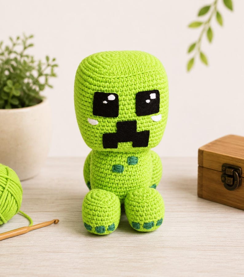 May include: A lime green crocheted plush toy featuring a large square head with black felt eyes and a black mouth. The body has small dark green square patches on the chest and feet, with white accents under the eyes, crafted in a yarn texture.