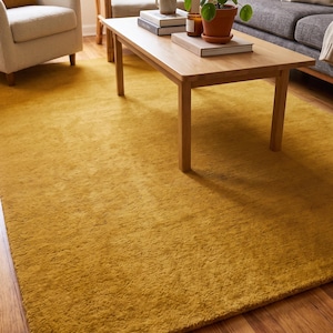 Close-up view of a handmade hand tufted mustard yellow wool area rug with a plush textured surface and neatly finished rolled edge revealing durable cotton canvas backing. Modern minimalist solid color rug crafted from premium New Zealand wool, ideal for living room, bedroom, office, nursery, luxury home decor, custom size area rug, contemporary interior, housewarming gift, and new home gift.