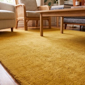 Close-up view of a handmade hand tufted mustard yellow wool area rug with a plush textured surface and neatly finished rolled edge revealing durable cotton canvas backing. Modern minimalist solid color rug crafted from premium New Zealand wool, ideal for living room, bedroom, office, nursery, luxury home decor, custom size area rug, contemporary interior, housewarming gift, and new home gift.