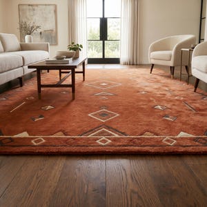 Hand tufted wool rug in rich terracotta featuring a southwestern tribal geometric pattern. Bold diamond medallion centre with concentric borders, ivory and charcoal accents, aztec-inspired motifs. Plush medium pile, artisan crafted, placed on warm oak hardwood floor. Perfect for boho living room, rustic bedroom or as a unique housewarming gift. Custom sizes available.
