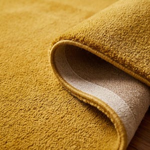 Close-up view of a handmade hand tufted mustard yellow wool area rug with a plush textured surface and neatly finished rolled edge revealing durable cotton canvas backing. Modern minimalist solid color rug crafted from premium New Zealand wool, ideal for living room, bedroom, office, nursery, luxury home decor, custom size area rug, contemporary interior, housewarming gift, and new home gift.