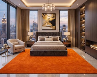 May include: A bedroom featuring a dark gray upholstered bed with neutral bedding, centered on a large vibrant orange area rug. The room includes a beige armchair, two bedside tables with lamps, a linear fireplace, and wall art with gold accents.