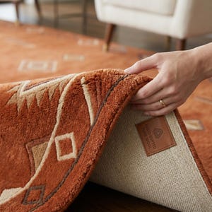 May include: A rust-coloured area rug with a geometric pattern in cream and brown. The rug has a thick, plush pile and a tan fabric backing. A brown leather label is stitched to the corner, featuring the text: HAND-TUFTED PURE WOOL.