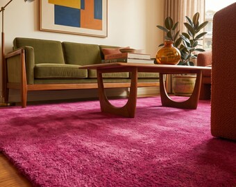 May include: A vibrant magenta area rug with a plush, textured pile covering the floor. The rug features a deep, saturated pink hue that provides a bold contrast to the olive green sofa, wooden coffee table, and abstract wall art in the room.